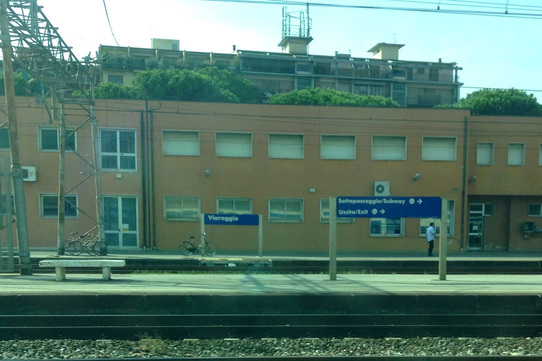 Il cartello della stazione di Viareggio sul binario, con l'edificio viaggiatori e i pini marittimi alle spalle